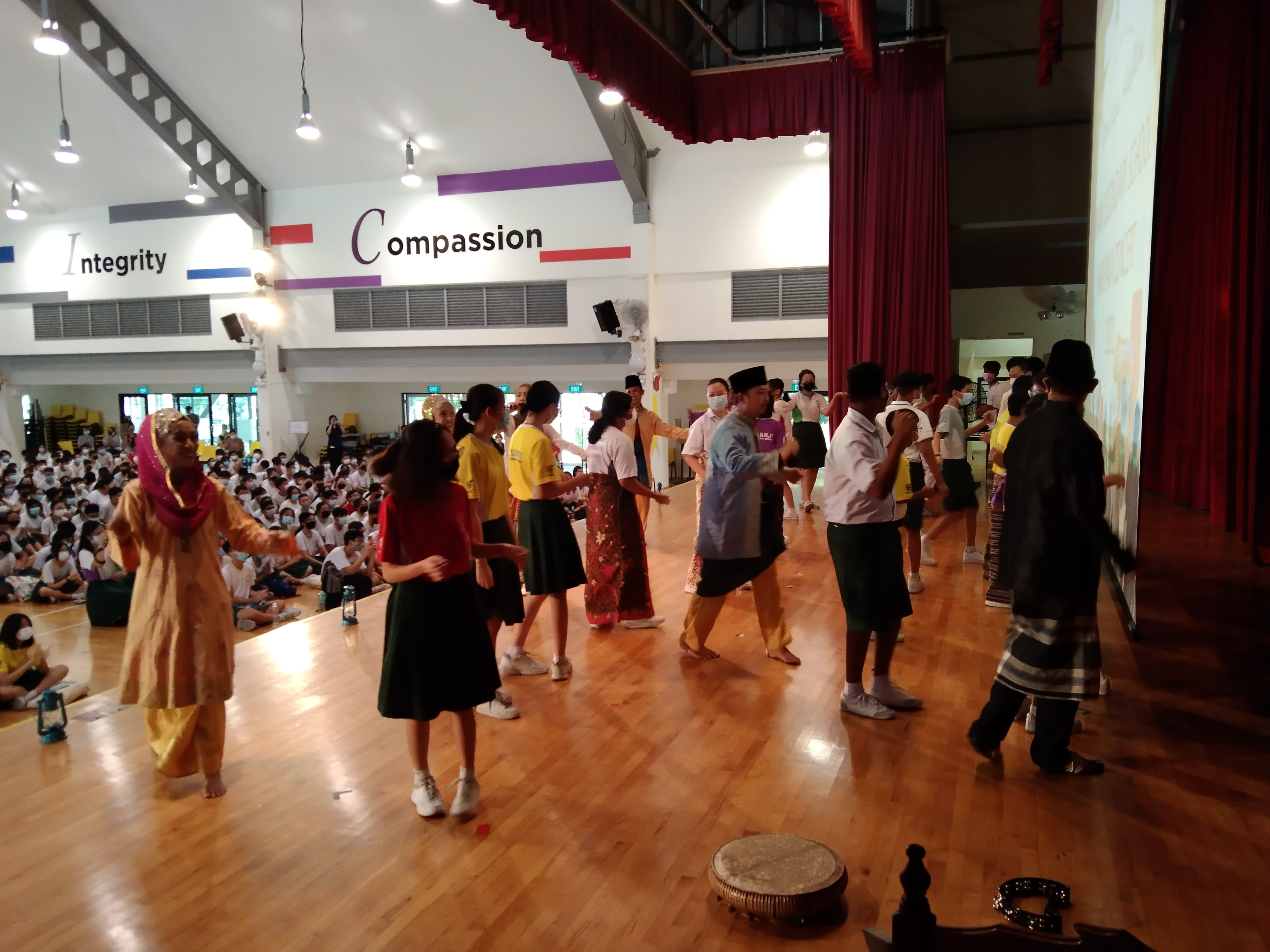 Students in traditional costumes perform a cultural dance in a school hall with "Integrity" and "Compassion" on the wall.